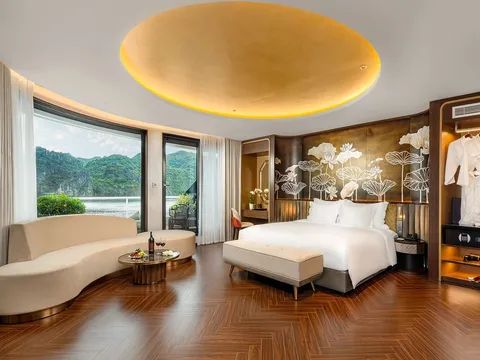 A luxury cruise cabin with a king bed, curved sofa, herringbone wood floor, gold circular ceiling feature, and large windows revealing karst limestone islands on the water outside.