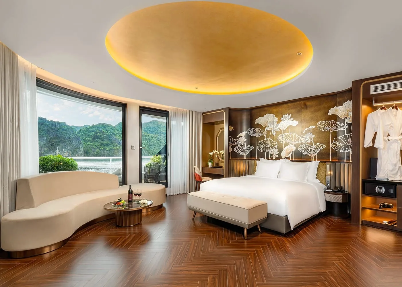A luxury cruise cabin with a king bed, curved sofa, herringbone wood floor, gold circular ceiling feature, and large windows revealing karst limestone islands on the water outside.