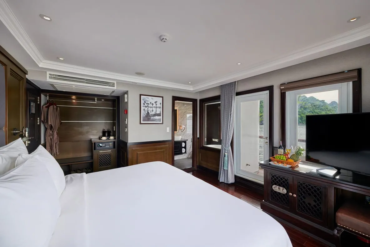 A luxury cruise cabin interior with a bed, dark wood furniture, television, and windows showing karst mountains and water outside.