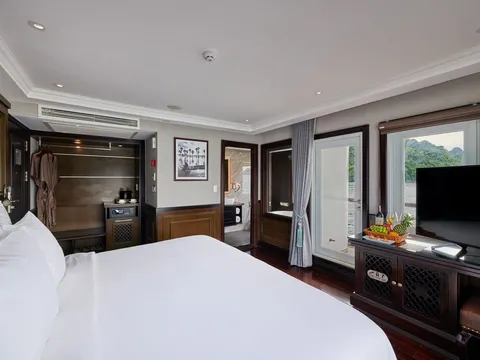A luxury cruise cabin interior with a bed, dark wood furniture, television, and windows showing karst mountains and water outside.
