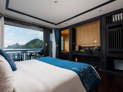 A boat cabin interior with a white bed, dark wood furnishings, and large windows showing limestone karst islands and water outside.