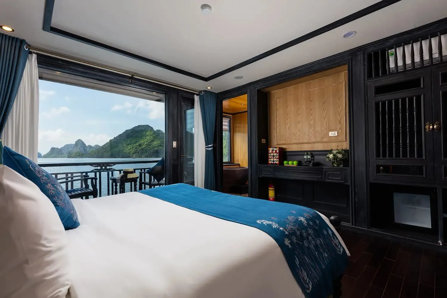 A boat cabin interior with a white bed, dark wood furnishings, and large windows showing limestone karst islands and water outside.