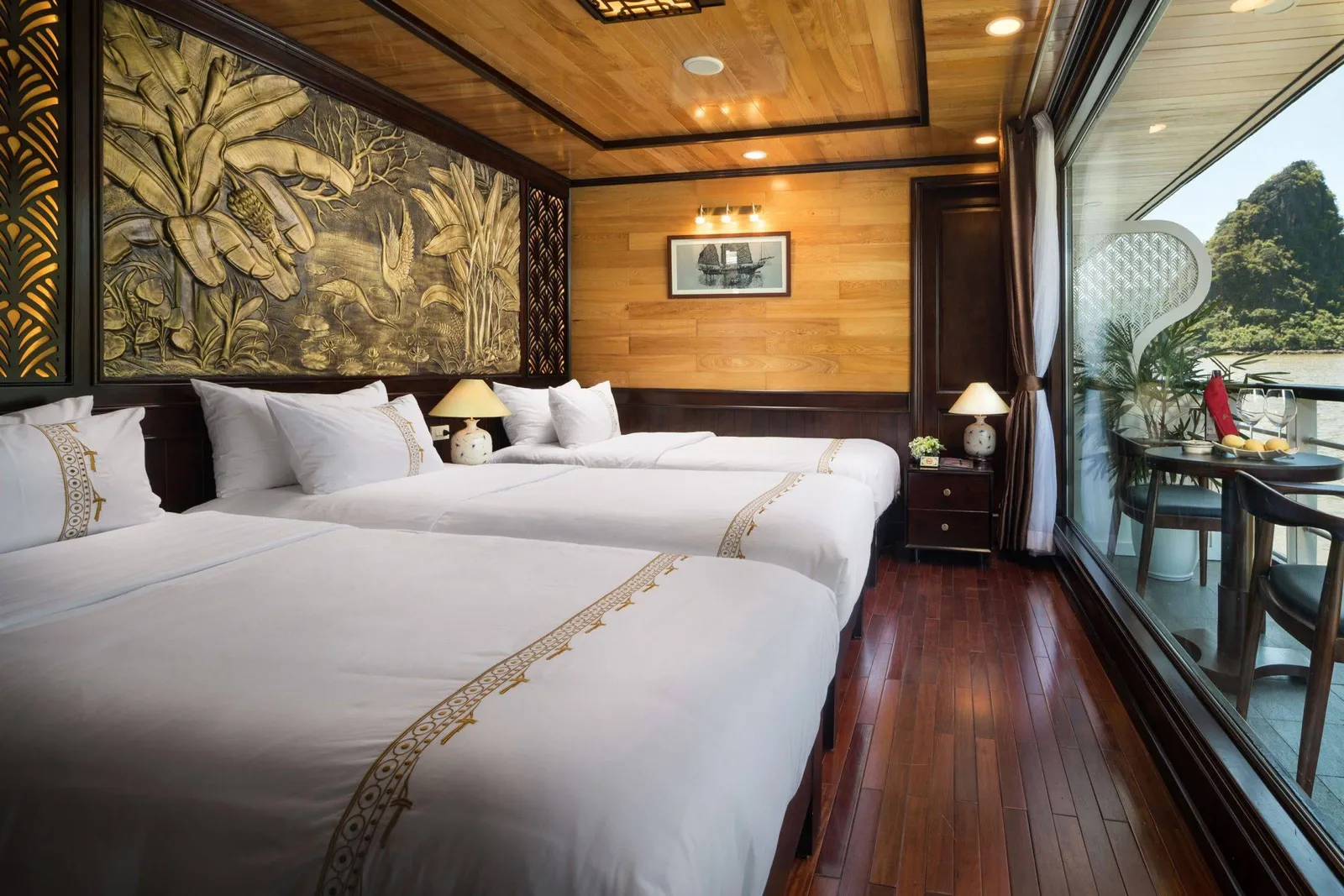 A wood-paneled cabin with three white-linen beds embroidered with gold trim, a carved banana-leaf relief on the wall, and a large window opening to a balcony with a table and a limestone karst island beyond.
