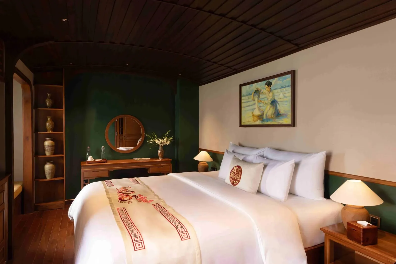 A cabin with a large white bed, a cream runner embroidered with a red dragon and key-pattern border, a red medallion cushion, dark wood curved ceiling, green walls, a wooden vanity with a round mirror, shelves of vases, and a framed painting on the wall.