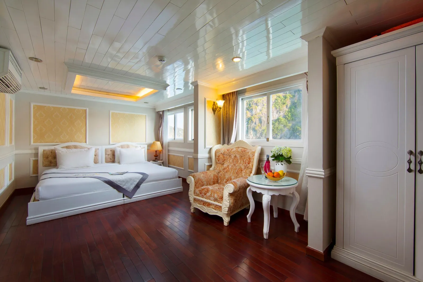 A cabin with a large white bed, an orange upholstered armchair, a small glass table with fruit and wine, a white wardrobe, and a window showing green limestone karst cliffs.