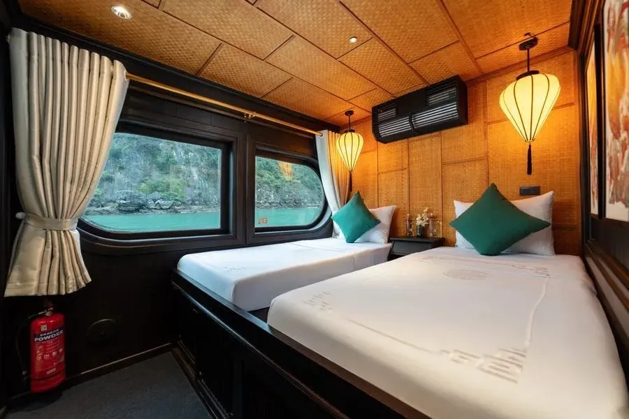 A twin-bed cabin with white linens, teal cushions, woven bamboo walls and ceiling, silk lanterns, and windows showing turquoise water and a forested limestone karst; a red fire extinguisher with printed label sits by the curtain, and a subtle pattern is embroidered on the bed covers.