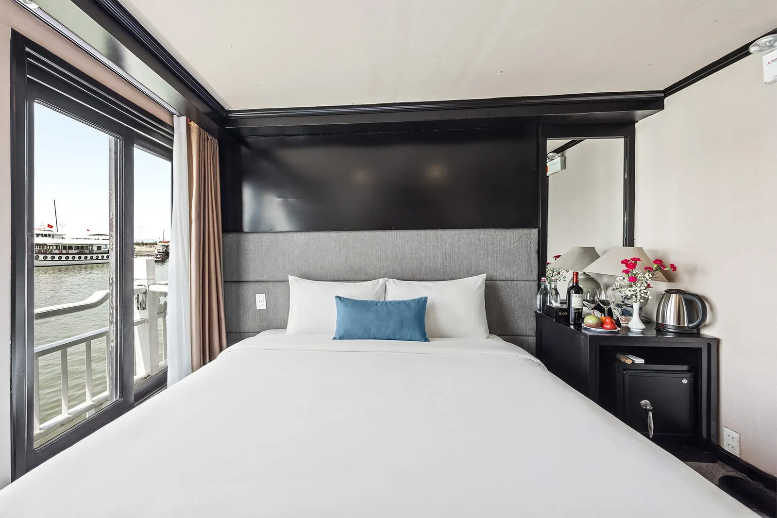 A cabin with a double bed, white linens and a blue cushion, a window and balcony railing overlooking water with another vessel, and a side table holding wine, fruit, flowers and a kettle.