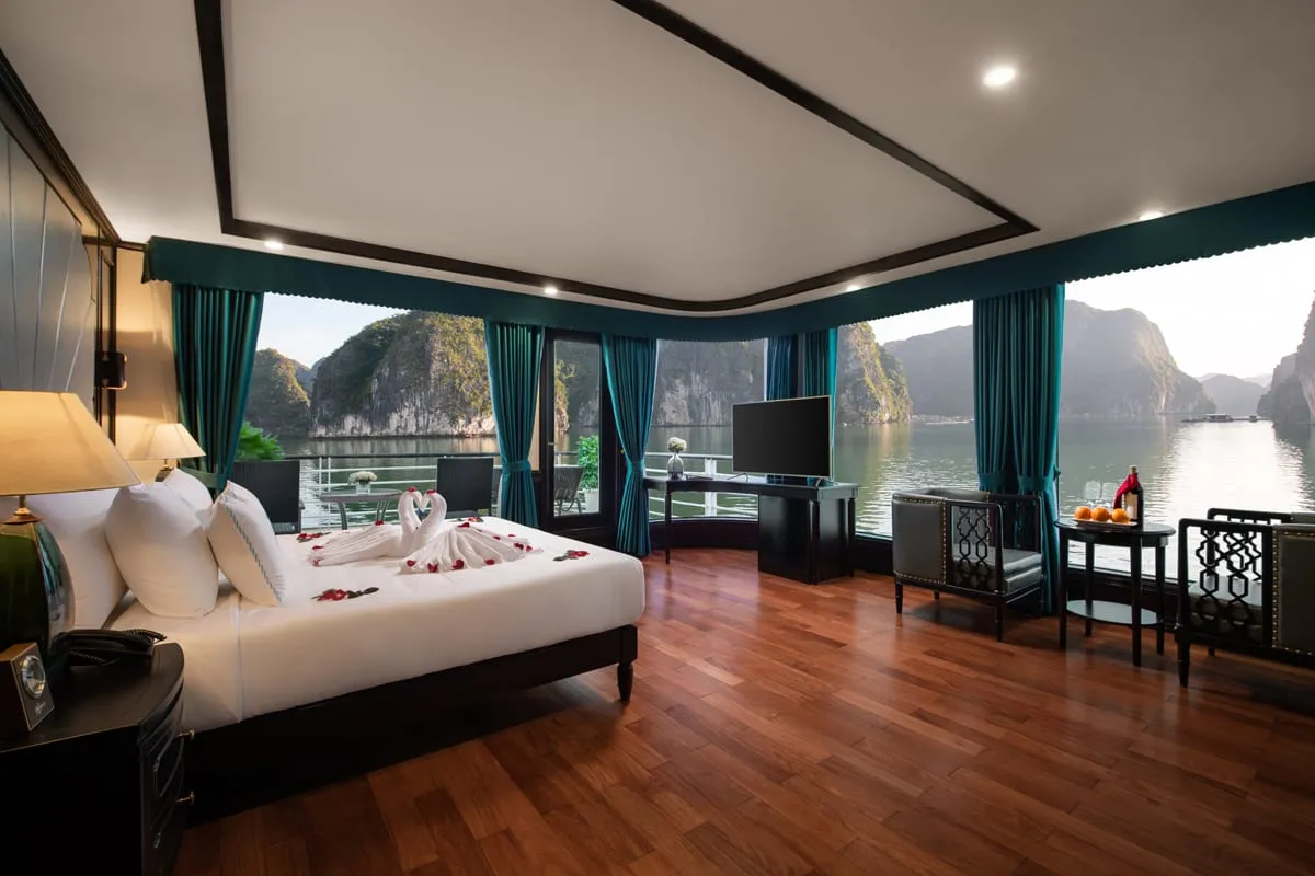 A luxury cabin interior with a decorated bed, wooden floors, teal curtains, and floor-to-ceiling windows overlooking limestone karsts and calm water.