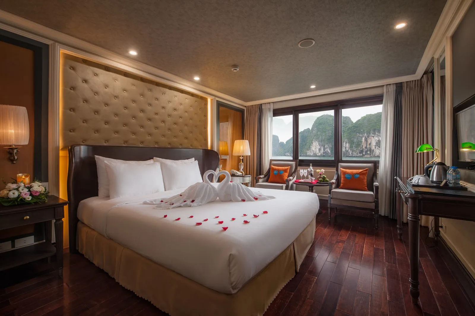 A cruise cabin with a double bed decorated with towel swans and red rose petals, with windows showing green limestone karst cliffs over the water.