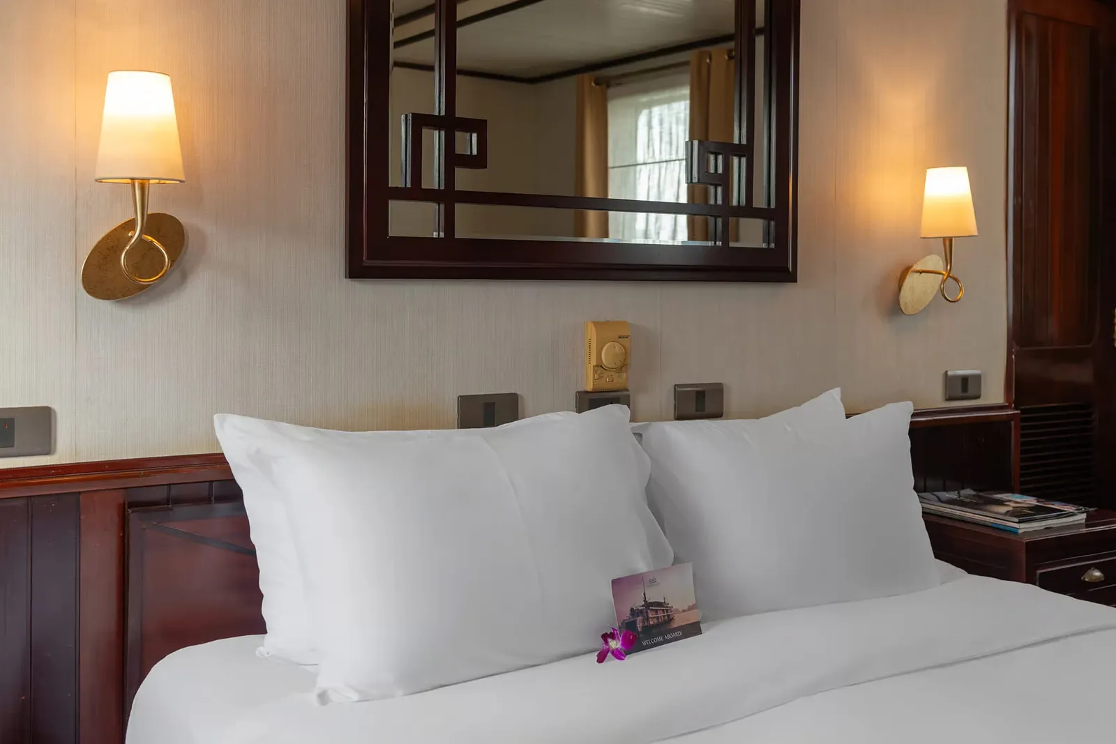 A cabin bed with white pillows and linens under a dark wood framed mirror, flanked by two wall lamps, with a 'Welcome Aboard' card showing a boat and a purple orchid on the bed.