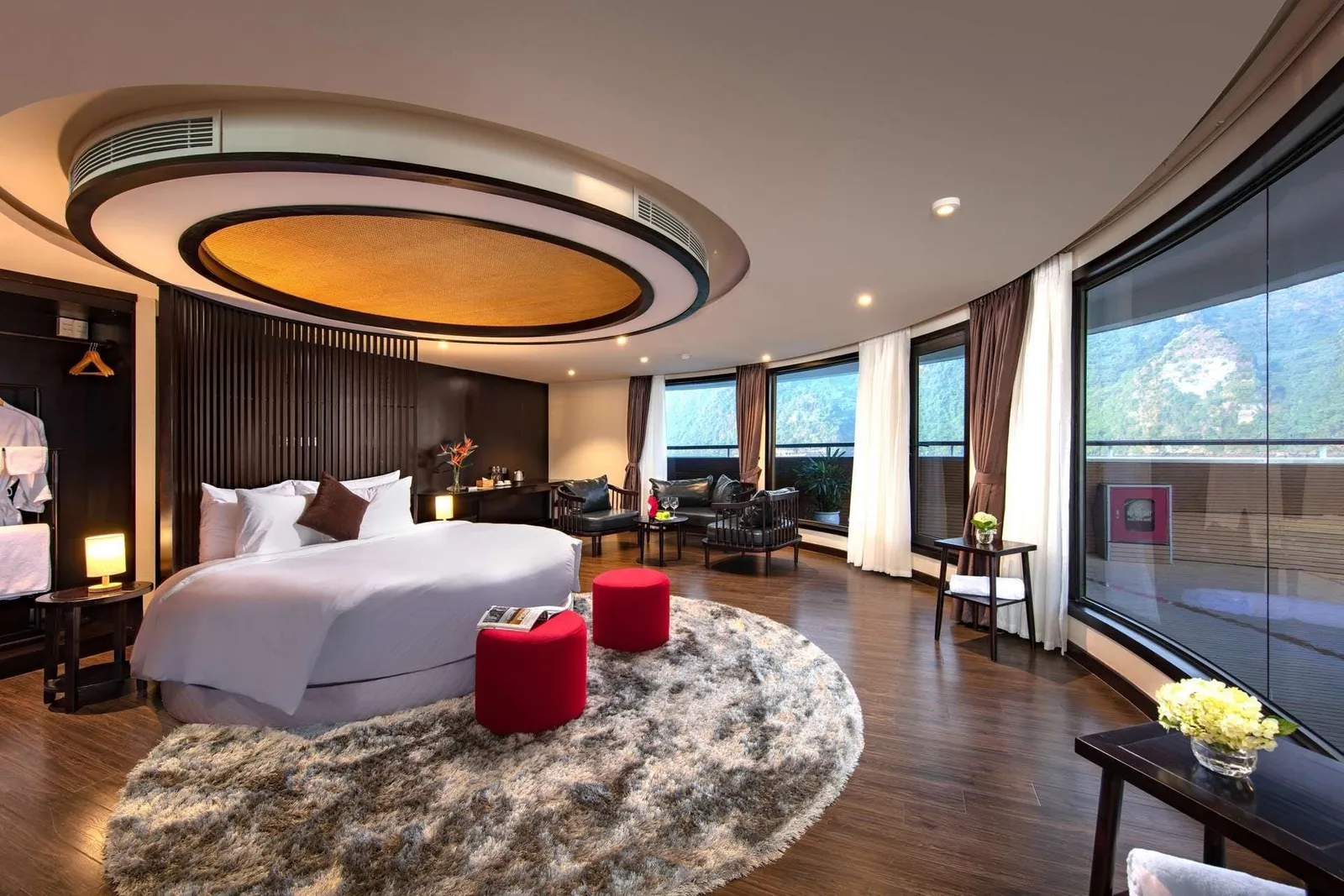 A spacious curved-wall suite with a round white bed, shaggy round rug, red stools, sitting area and floor-to-ceiling windows overlooking forested limestone hills and a balcony.