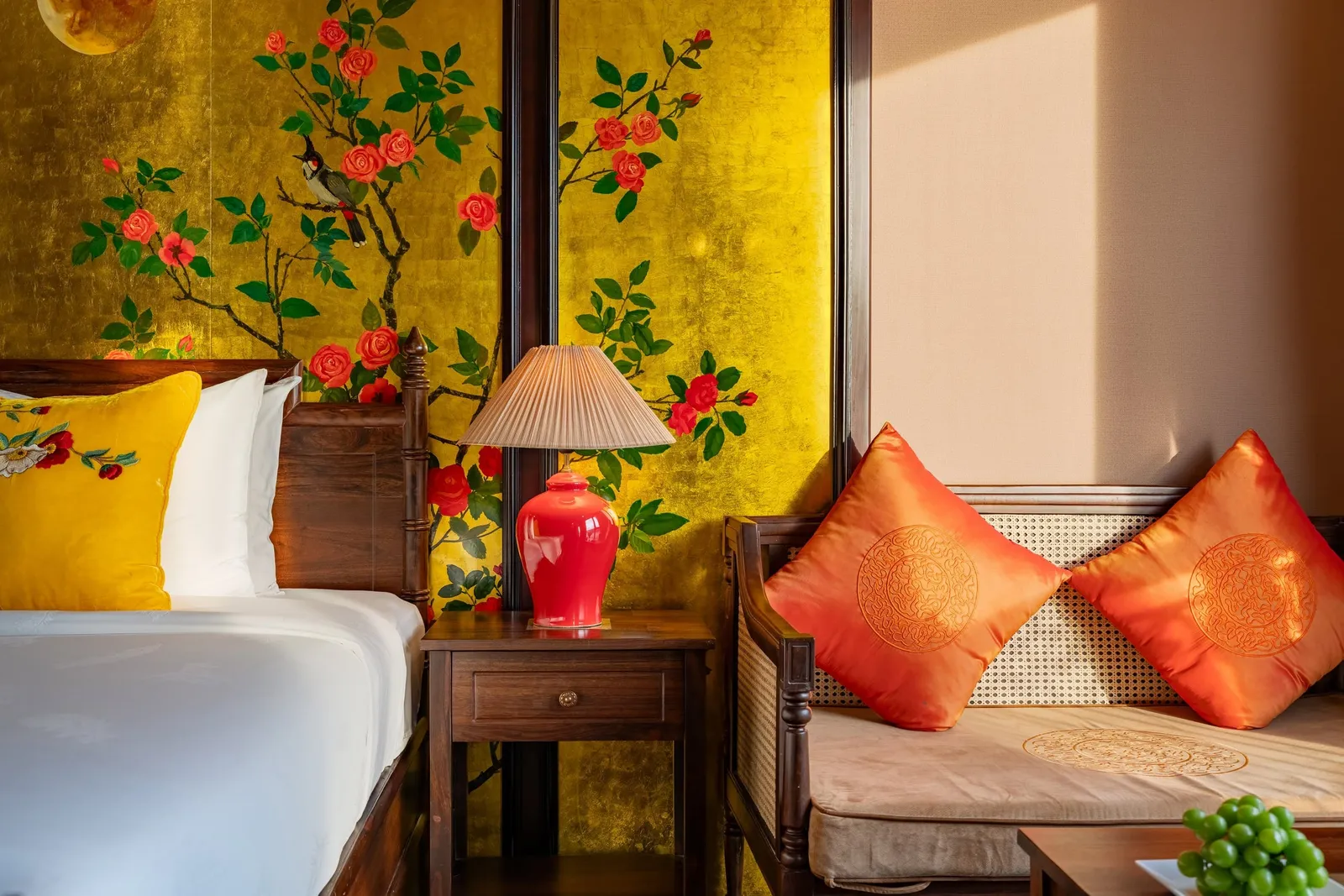 Bed against a hand-painted floral panel, a red lamp on the side table and orange cushions on the daybed