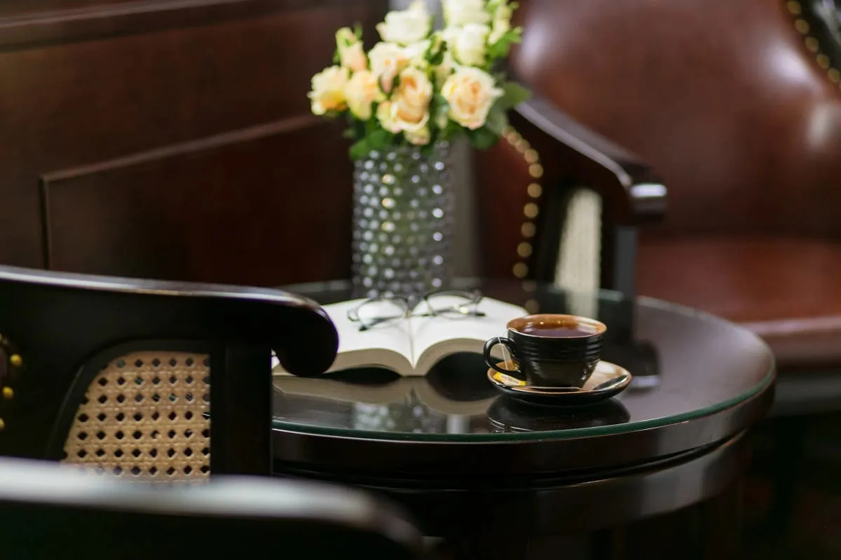 An interior cabin scene with a tea cup on a round glass table, an open book, flowers in a vase, and dark wood furniture including a chair with cane backing.