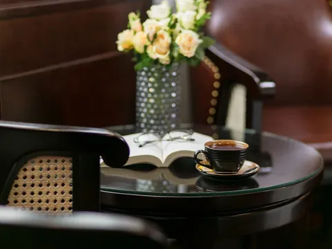 An interior cabin scene with a tea cup on a round glass table, an open book, flowers in a vase, and dark wood furniture including a chair with cane backing.