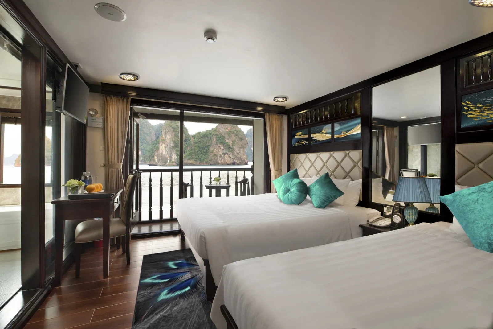 A cabin interior with two beds, turquoise pillows, dark wood trim, and a window showing limestone karst mountains typical of Ha Long Bay.