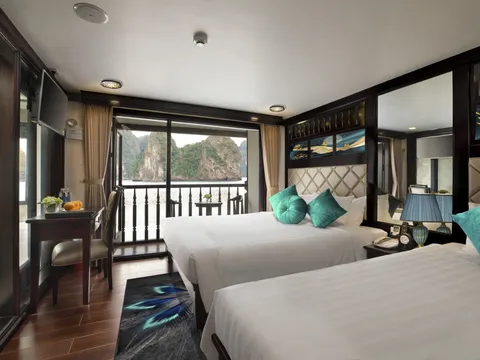 A cabin interior with two beds, turquoise pillows, dark wood trim, and a window showing limestone karst mountains typical of Ha Long Bay.