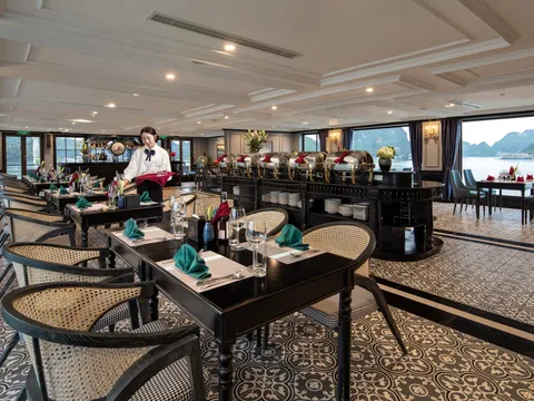 An elegant dining room interior with set tables, wicker chairs, ornate ceiling, and large windows showing karst mountains and water outside.