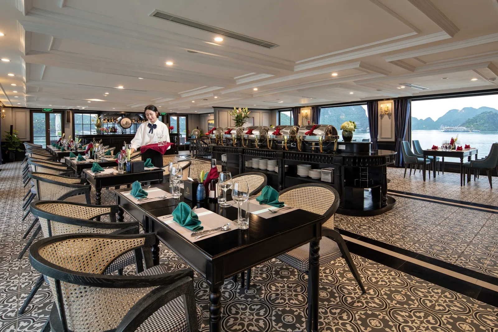 An elegant dining room interior with set tables, wicker chairs, ornate ceiling, and large windows showing karst mountains and water outside.