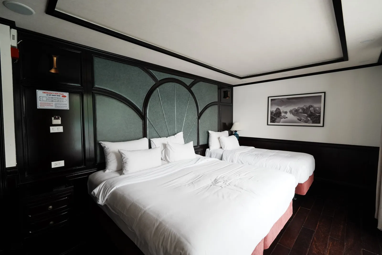 A cabin with dark wood paneling and green padded headboard, holding two white-linen beds with pink bases, a framed black-and-white Ha Long Bay karst photo on the wall, and an emergency exit plan posted by the door.