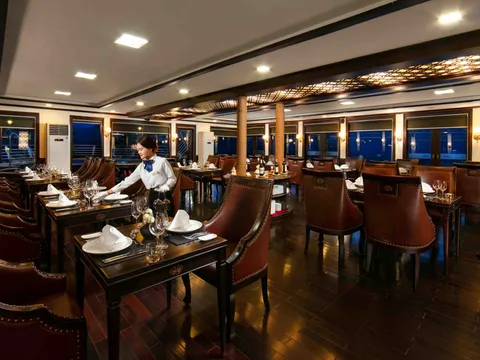 An elegant dining room with set tables, brown leather chairs, large windows showing darkness outside, and a staff member in uniform.