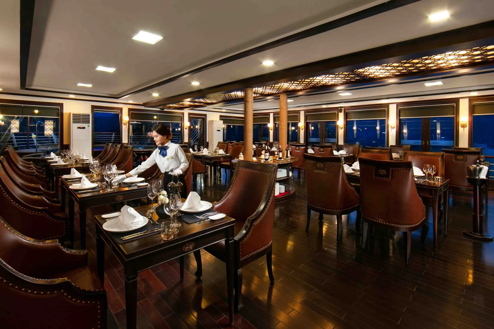 An elegant dining room with set tables, brown leather chairs, large windows showing darkness outside, and a staff member in uniform.