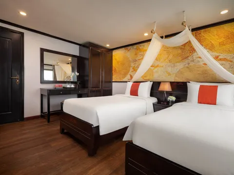 A cruise cabin interior with two beds, white canopy draping, orange accent pillows, dark wood furniture, and a golden marble feature wall.