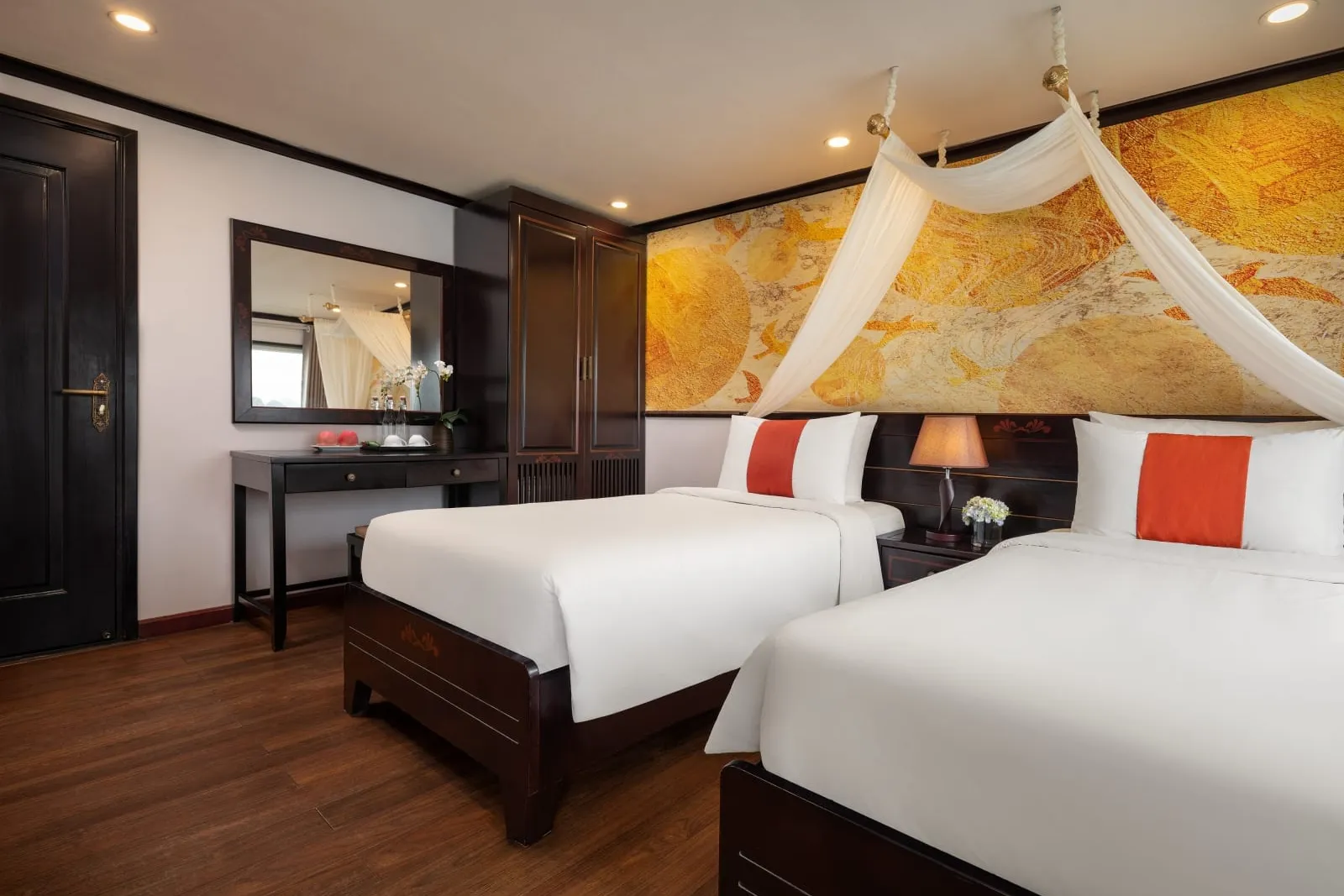 A cruise cabin interior with two beds, white canopy draping, orange accent pillows, dark wood furniture, and a golden marble feature wall.