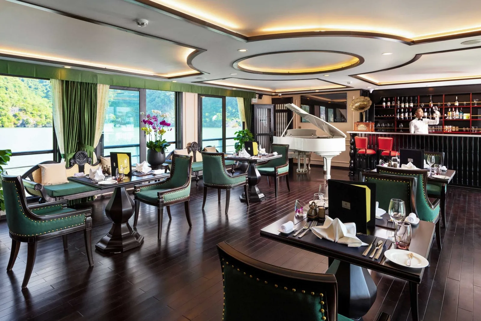 An upscale dining room and bar area on a cruise boat with green velvet chairs, dark wood floors, a white grand piano, and large windows showing karst limestone mountains and water outside.