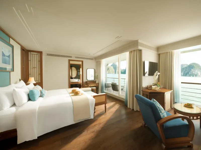 A luxury cabin interior with a bed, seating area, and large windows showing limestone karst formations and water outside.