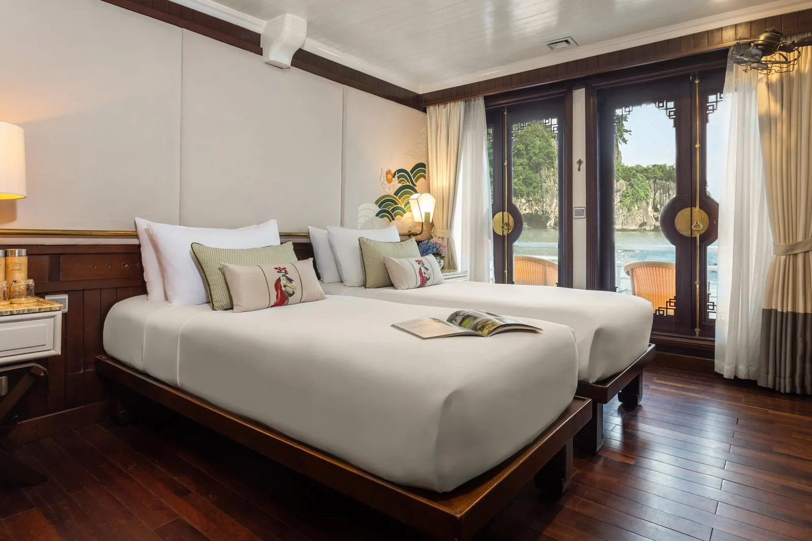 A luxury cruise cabin with two beds, dark wood furniture, and glass doors opening to a view of limestone karsts and water.