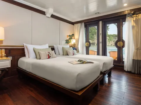 A luxury cruise cabin with two beds, dark wood furniture, and glass doors opening to a view of limestone karsts and water.