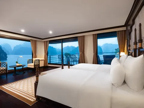 A well-appointed twin-bed cabin with dark wood four-poster beds, patterned rug, and large windows opening onto a balcony with karst limestone islands visible in the blue twilight.