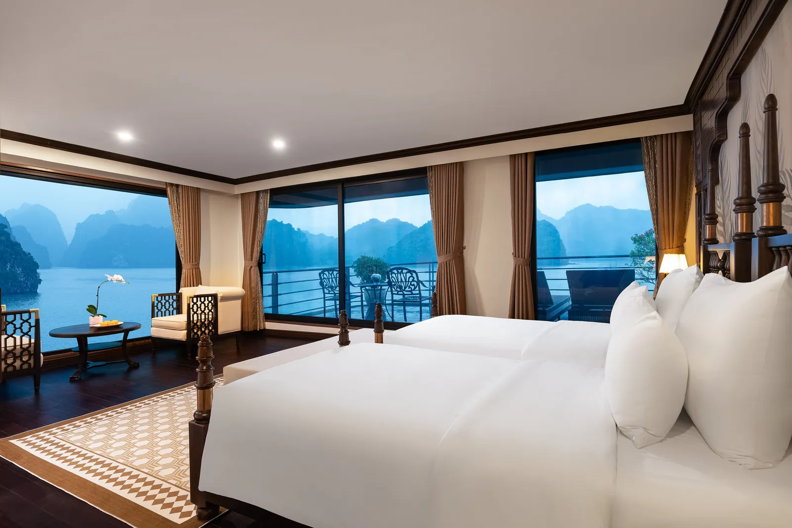 A well-appointed twin-bed cabin with dark wood four-poster beds, patterned rug, and large windows opening onto a balcony with karst limestone islands visible in the blue twilight.