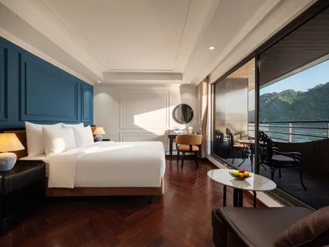 A luxury boat cabin with white bedding, dark wood floors, a vanity area, and sliding doors opening to a private balcony overlooking forested limestone karst islands and calm green water.