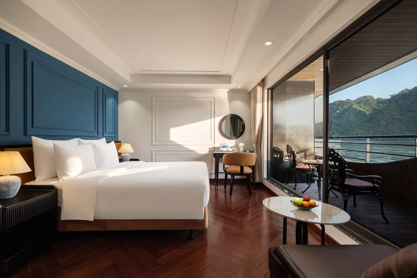 A luxury boat cabin with white bedding, dark wood floors, a vanity area, and sliding doors opening to a private balcony overlooking forested limestone karst islands and calm green water.