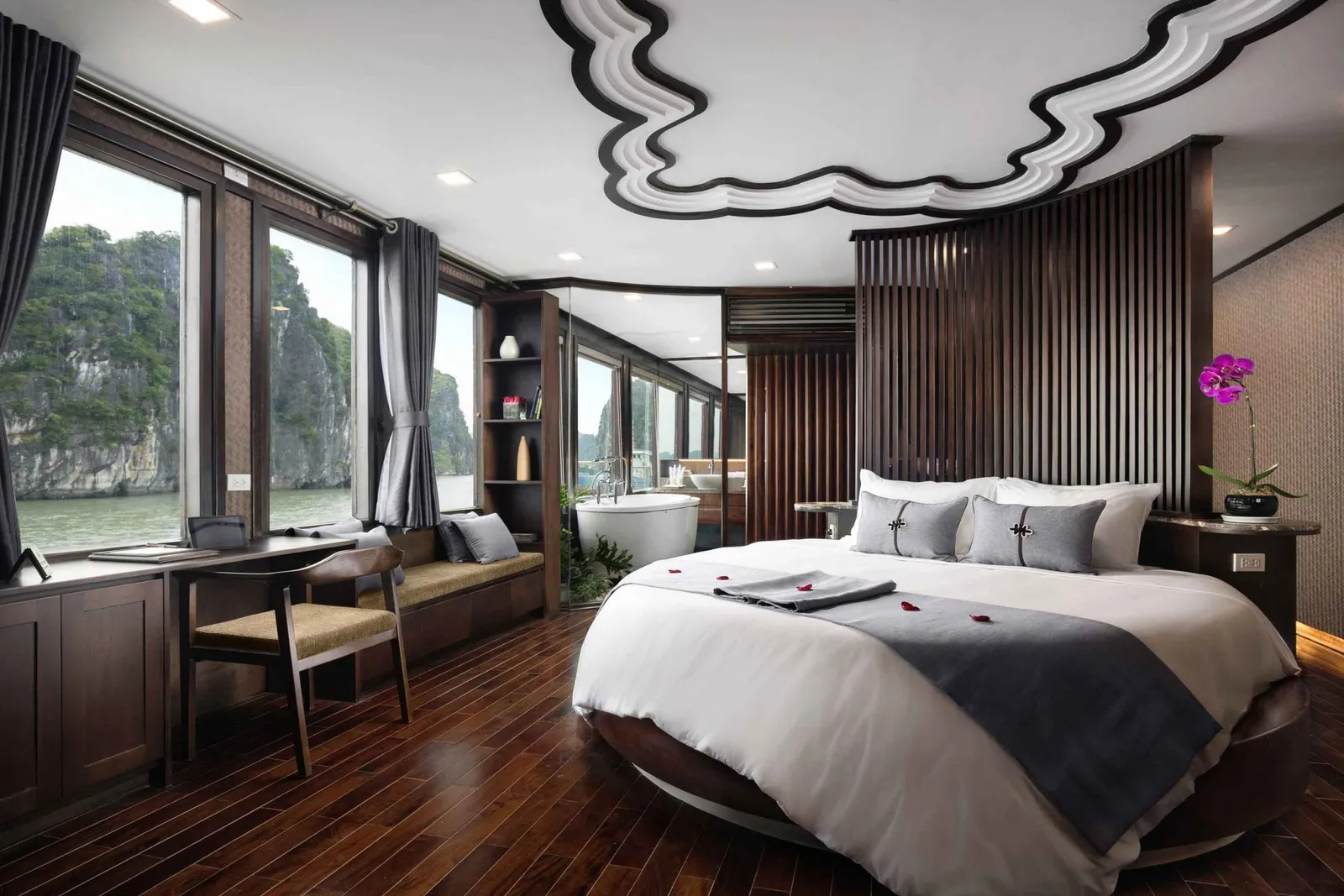 A luxury cabin interior with a made bed, wooden floors, large windows showing limestone karsts and water outside.