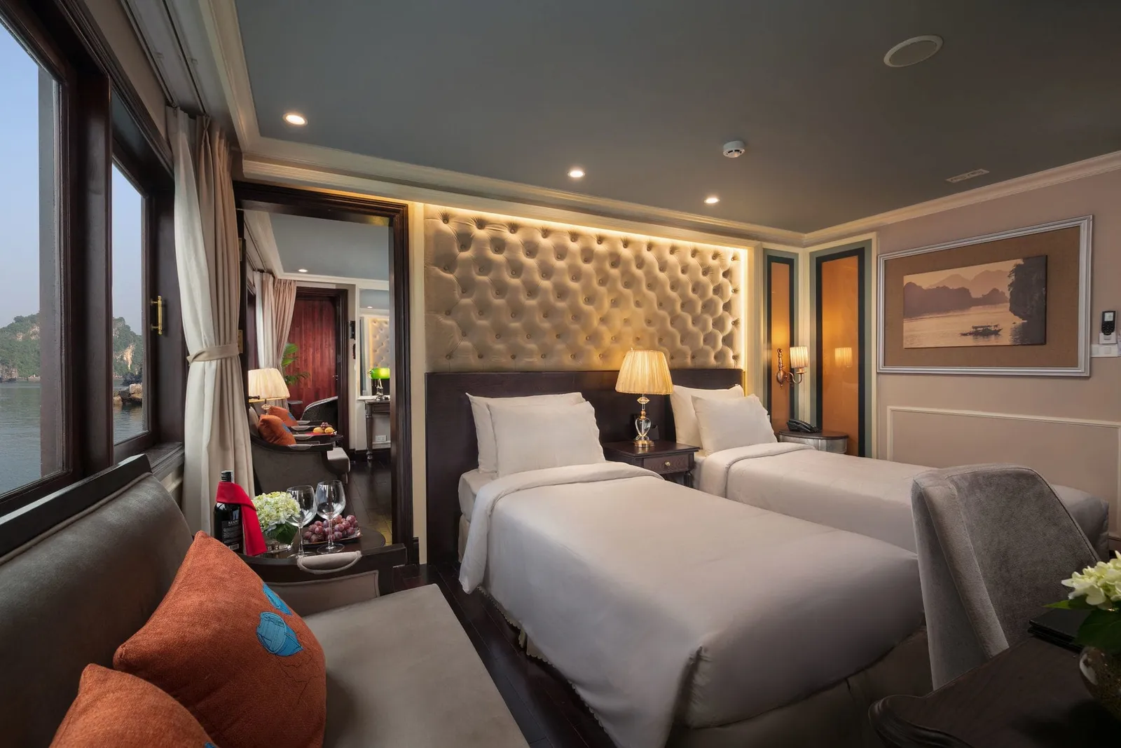 A cabin with two twin beds, a tufted headboard, lamps and a sofa with orange cushions, with windows showing calm water and a limestone karst.
