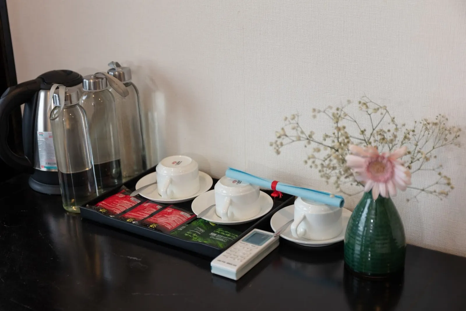 A dark tabletop against a cream wall holds an electric kettle, three glass water bottles, a tray with three upturned cups on saucers and tea sachets, an air-conditioner remote, and a small green vase with a pink gerbera and baby's breath; the cups carry a small printed logo.