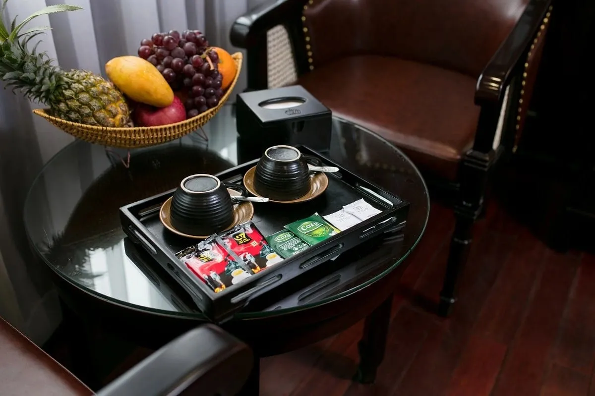 A round glass-topped table with a fruit basket, two black coffee cups on a tray with coffee and tea sachets, and leather armchairs on a dark wooden floor in an interior room.