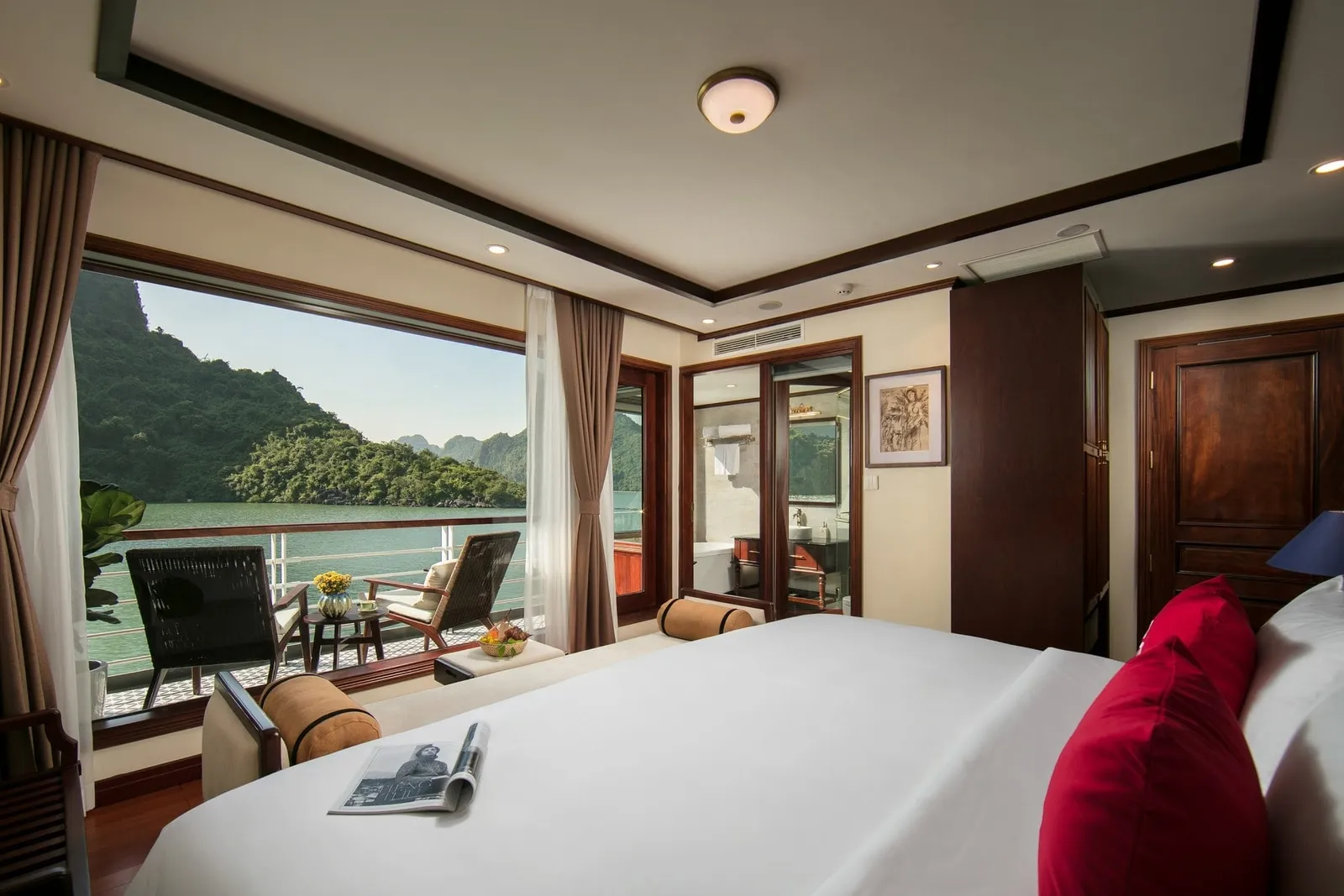 A luxury cabin interior with a white bed, private balcony with chairs, and limestone karst mountains visible across green water through the open doors.