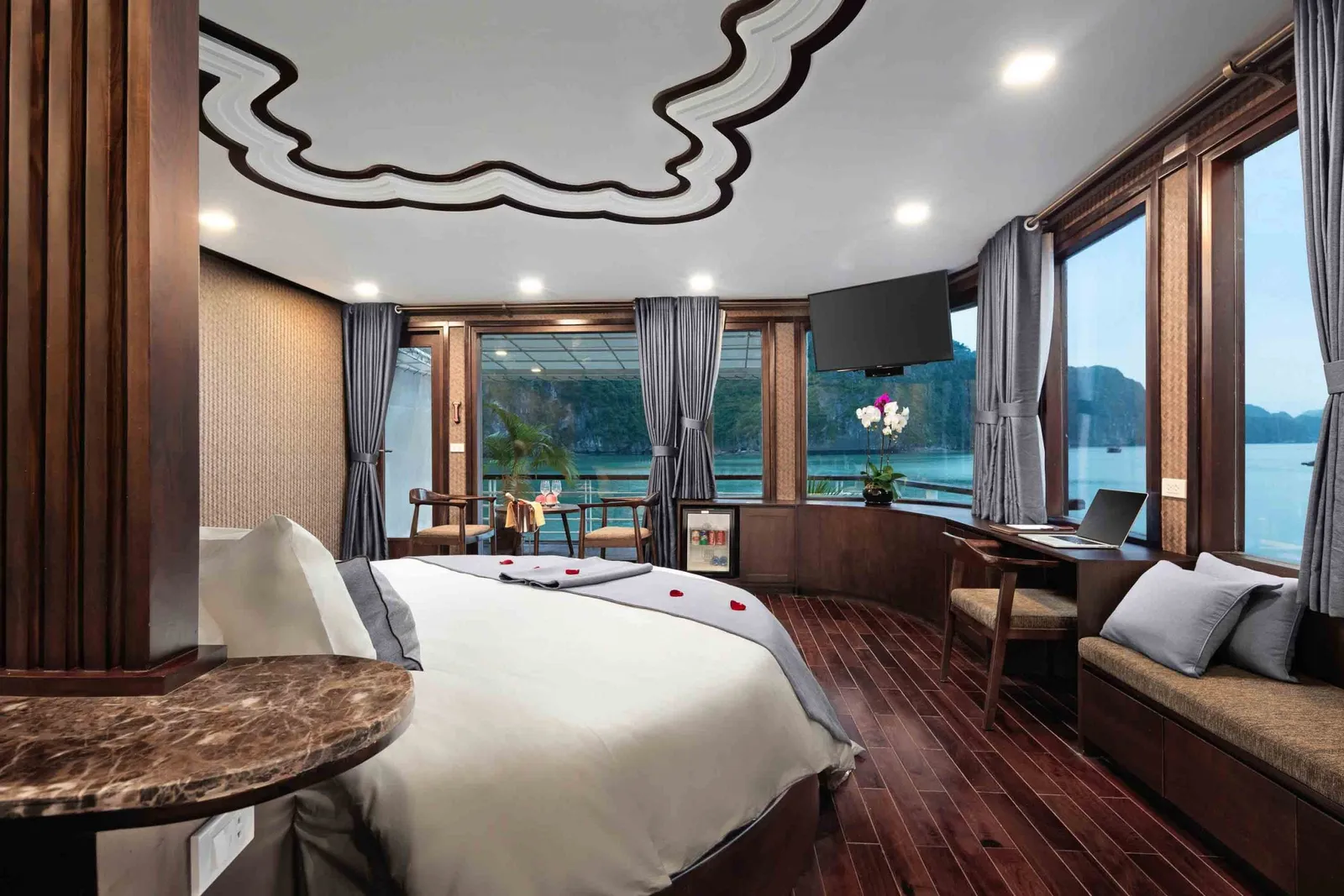 A boat cabin interior with a large bed, wooden furniture, and panoramic windows showing limestone karsts and water typical of Ha Long Bay.