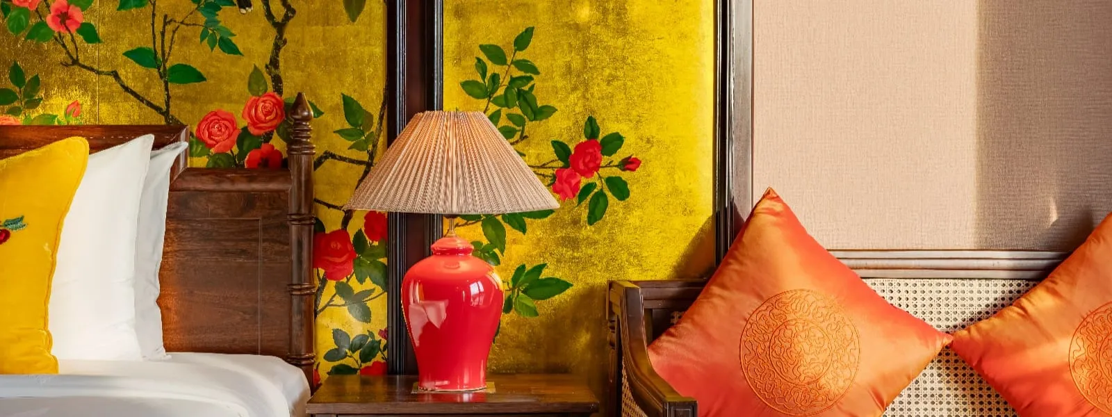 Cabin interior with a bed with white and yellow embroidered pillows, a red ceramic lamp on a wooden nightstand, gold-leaf floral wallpaper, and a rattan chair with two orange silk cushions.