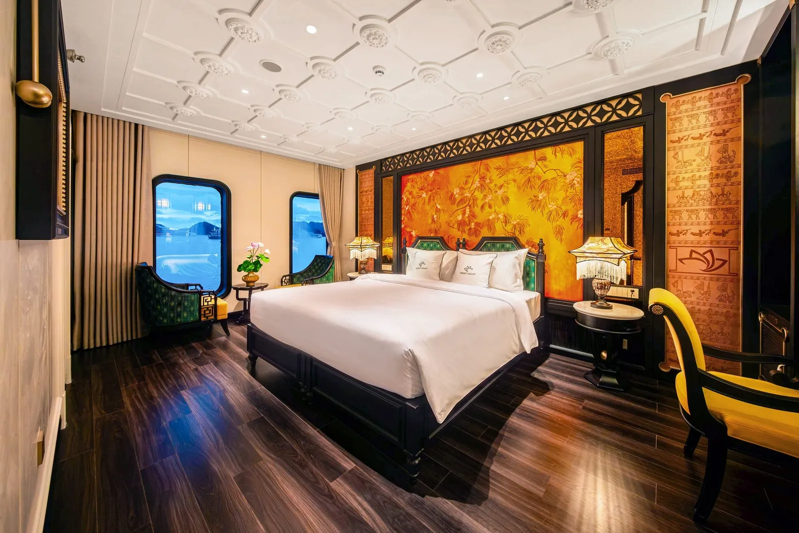 A luxurious cabin with a large white-linen bed, dark wood floor, orange lacquer-style wall panel, green and yellow chairs, and rounded windows showing blue water and limestone karsts, with small logos on the pillows.
