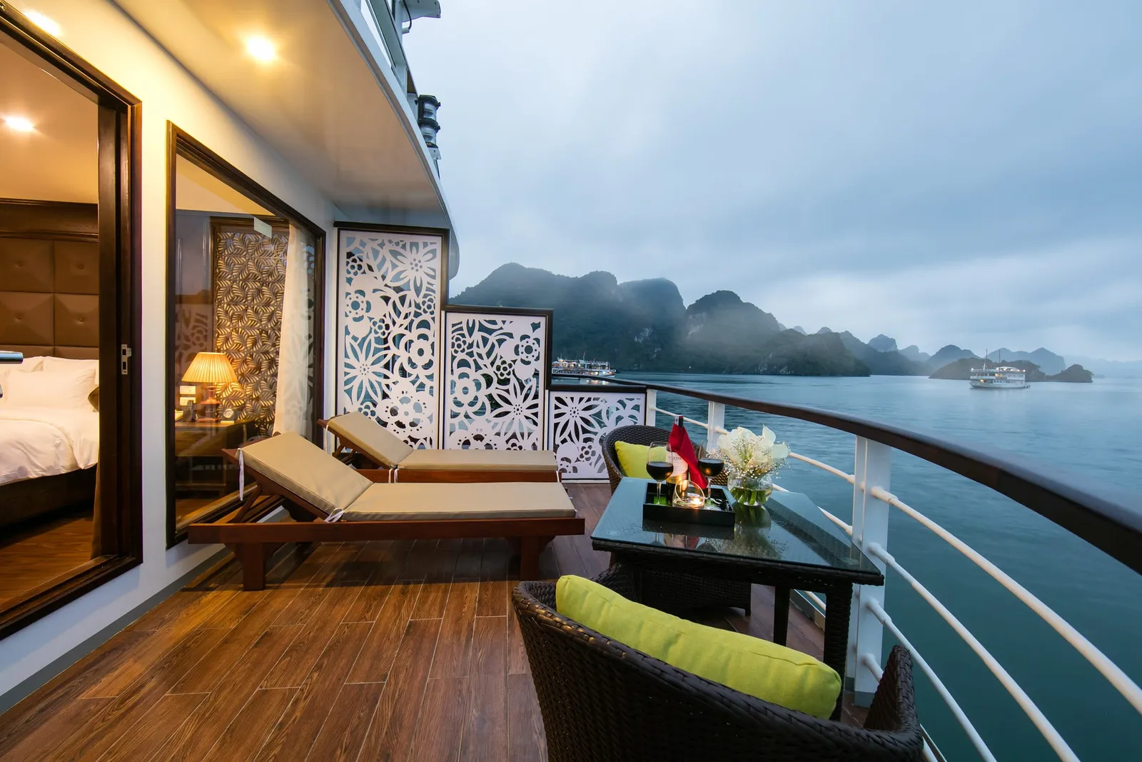 Private balcony with a sun lounger behind a carved screen, the cabin bed visible through the open door, karst in the haze