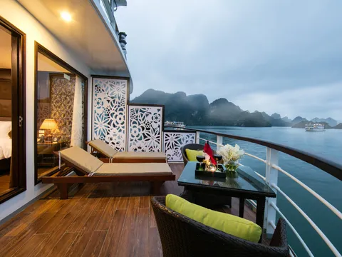 Private balcony with a sun lounger behind a carved screen, the cabin bed visible through the open door, karst in the haze