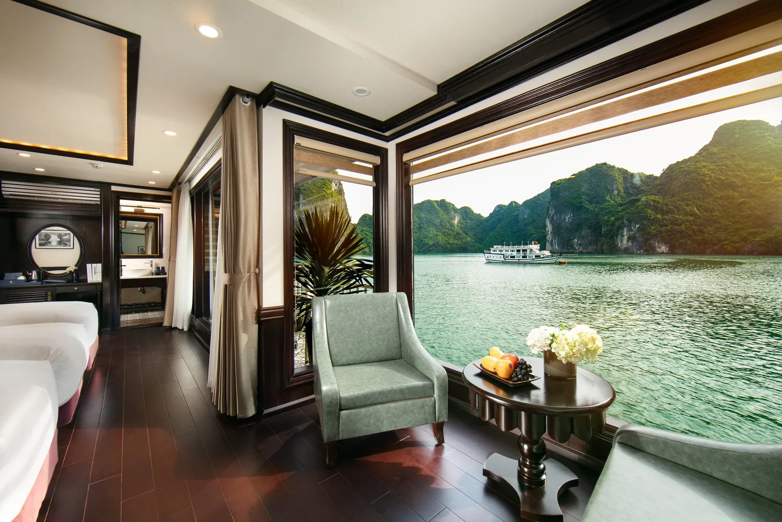 A cabin interior with beds, a dark wood floor, green armchairs and a round table with fruit and flowers, looking through a large window at green limestone karsts and a distant white ship on calm water.