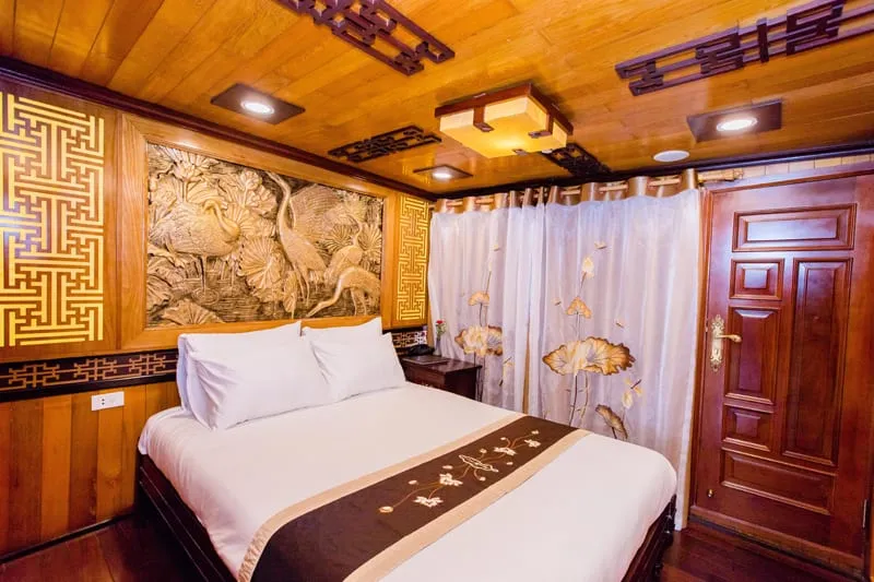 A cruise boat cabin interior with a double bed, carved wood wall art depicting cranes, decorative wooden ceiling panels with recessed lighting, embroidered curtains with lotus motifs, and a dark wood door.
