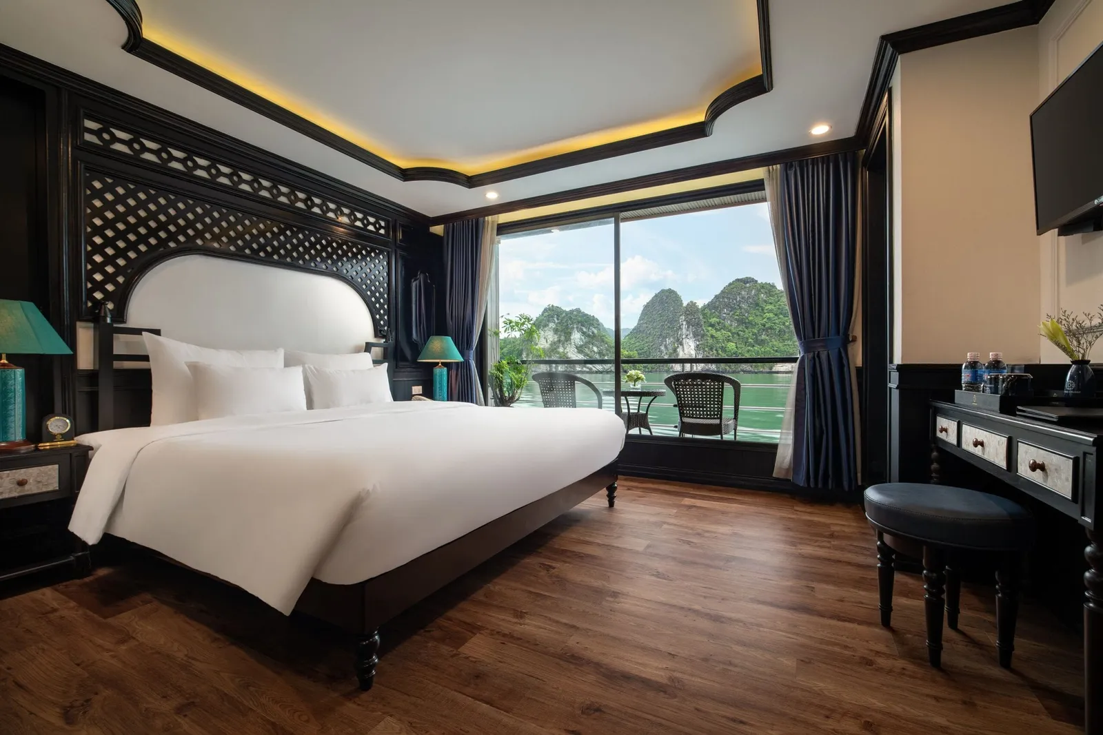 A luxury cabin interior with a white bed, dark wood furnishings, and a balcony view of dramatic limestone karsts rising from green water.