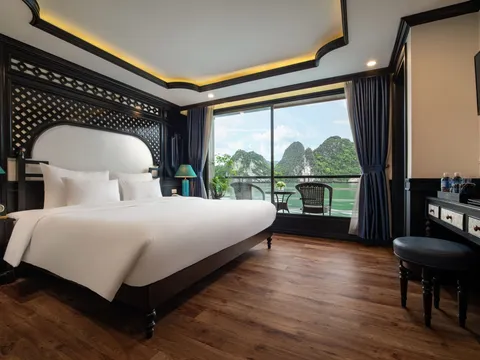 A luxury cabin interior with a white bed, dark wood furnishings, and a balcony view of dramatic limestone karsts rising from green water.