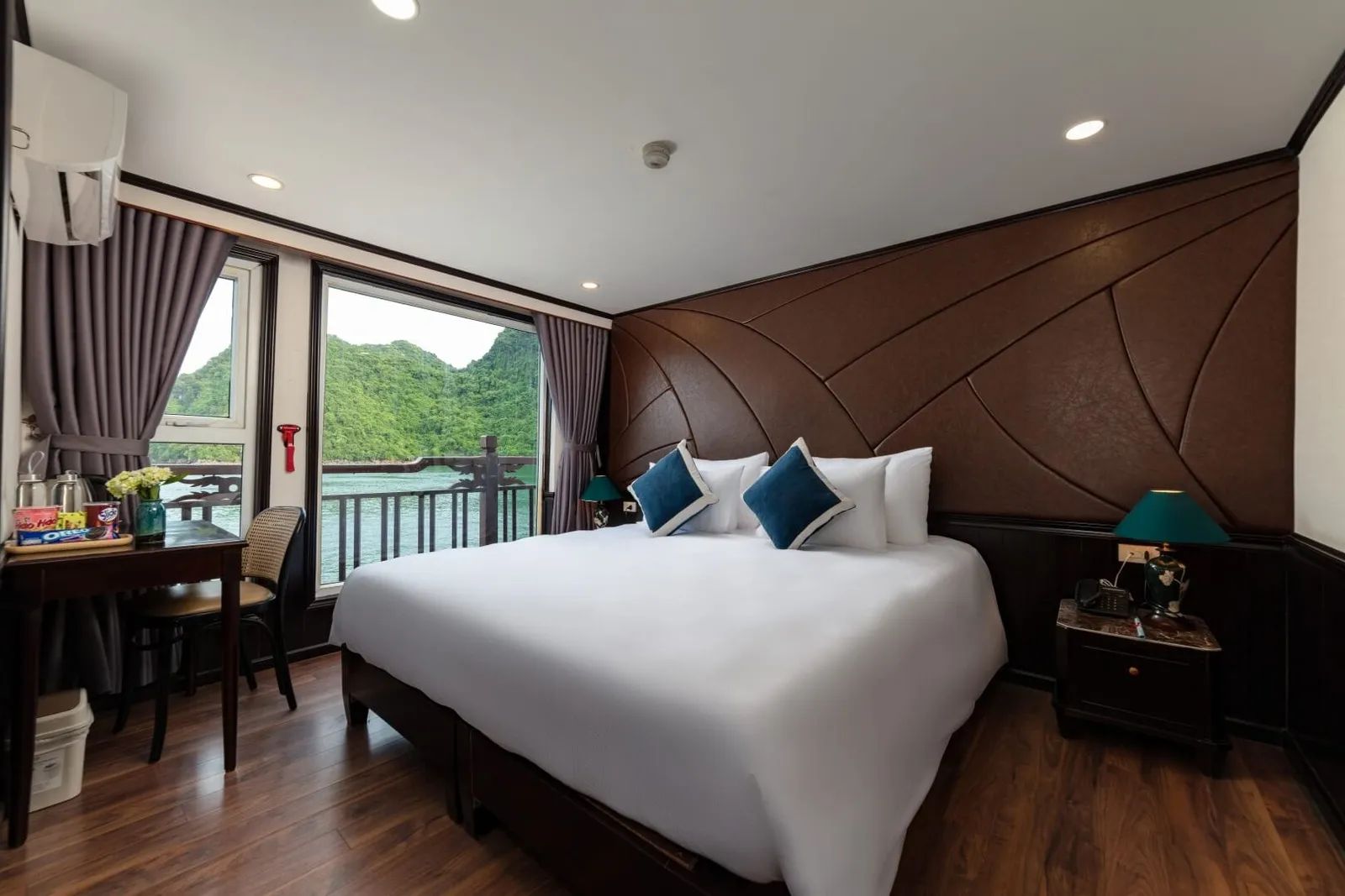 A cruise cabin with a large white double bed, brown leather-paneled headboard wall, wooden desk and bedside table, and a window and balcony railing overlooking green karst hills and water.